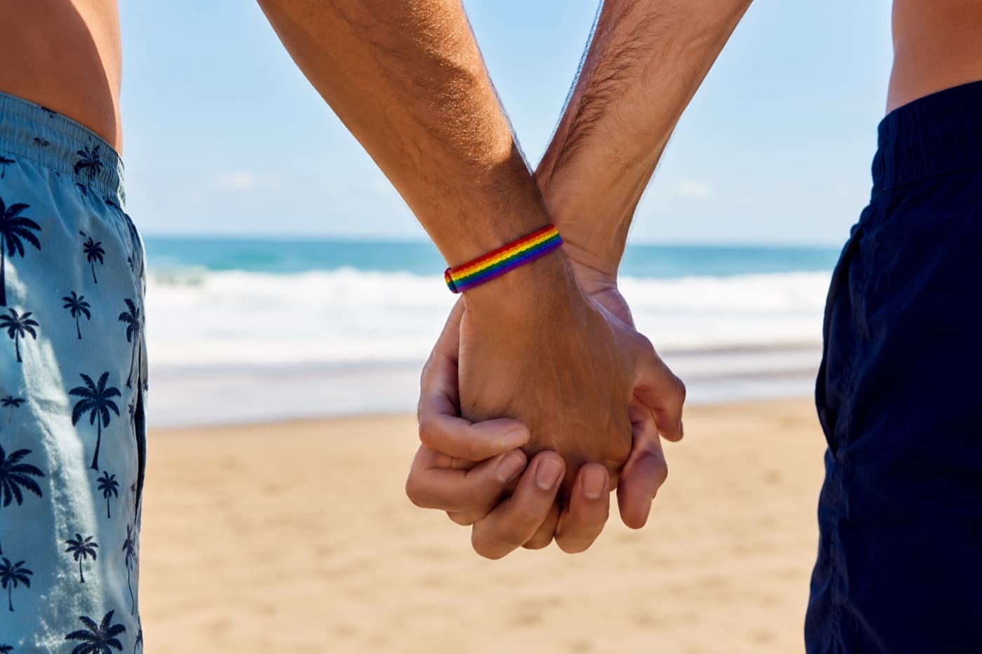 Two men holding hands on a beach, one wearing a rainbow wristband